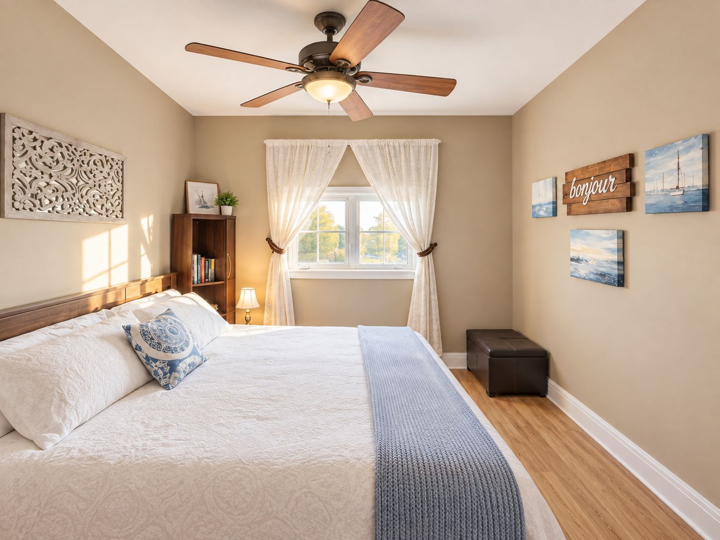Une chambre à coucher comprenant un lit, un ventilateur de plafond, une fenêtre avec des rideaux, une table de chevet avec une lampe, une commode et des œuvres d’art mural.