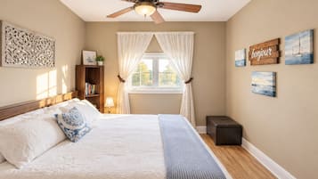 Une chambre à coucher comprenant un lit, un ventilateur de plafond, une fenêtre avec des rideaux, une table de chevet avec une lampe, une commode et des œuvres d’art mural.