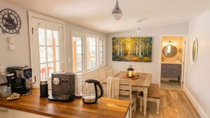 Une cuisine équipée d’une machine à café, d’un grille-pain et d’une bouilloire sur le plan de travail, une table à manger avec des chaises, un tableau représentant une forêt, et une salle de bain avec un miroir et un lavabo.