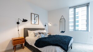 A bedroom with a bed, bedside lamps, a nightstand, a wall-mounted mirror, and a window. - 2BR | Gym, Pool | Bellevue | Sophari Towers I (Bellevue)
