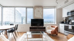 A modern living room with a large window, a flat-screen TV, a dining area, and a kitchen. - 2BR | Gym, Pool | Bellevue | Sophari Towers I (Bellevue)