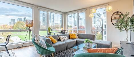 A modern living room with a grey sectional sofa, teal armchairs, and a glass coffee table. Large windows offer a view of the cityscape.