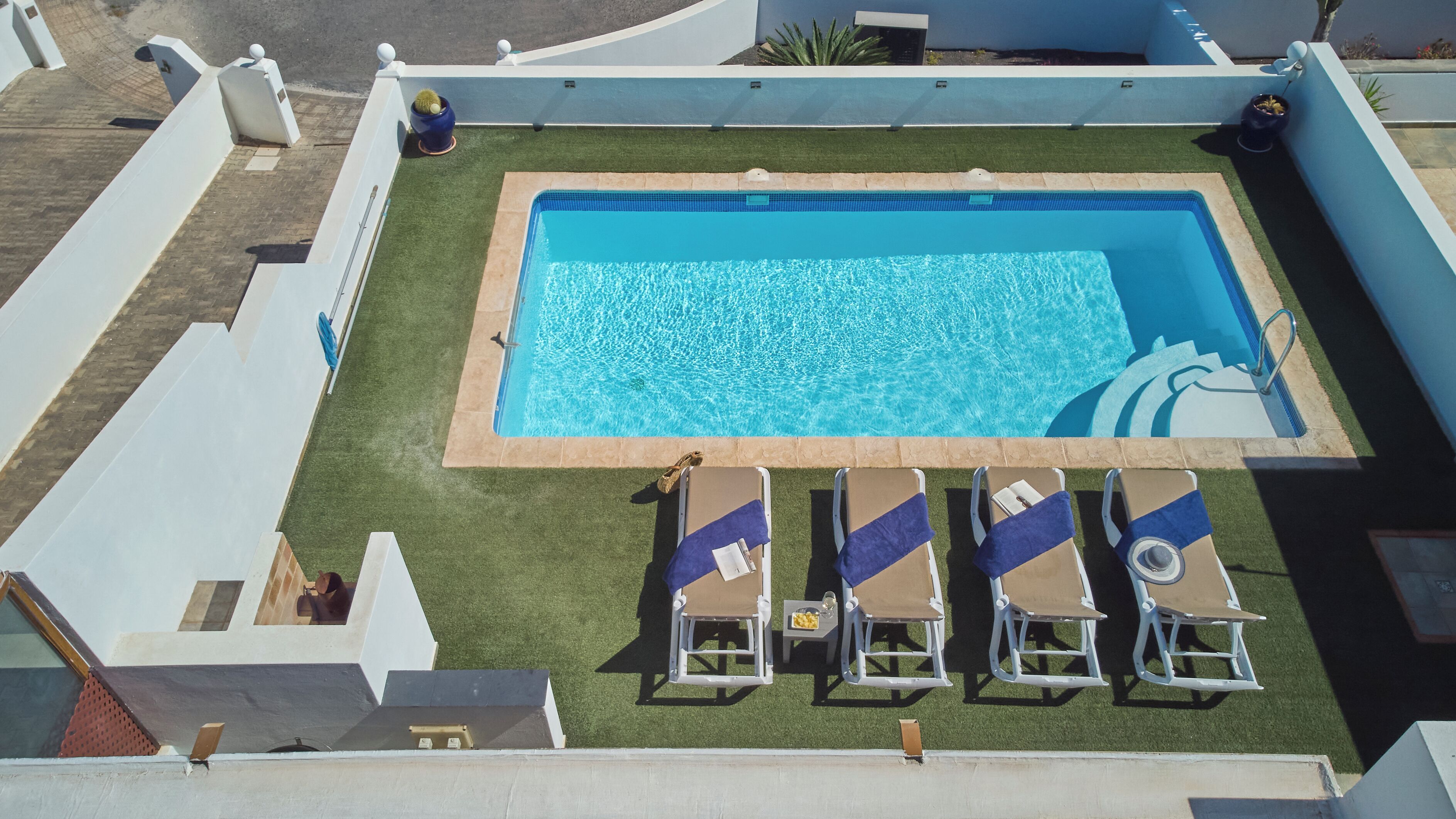 Vista aérea de una piscina rectangular con interior azul, rodeada por un solárium blanco. Hay cuatro sillas de descanso con toallas azules y una pequeña mesa con un cuenco de fruta.