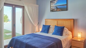 Un dormitorio con cama, mesita de noche con lámpara, un cuadro de un paisaje y una puerta corrediza de vidrio a un balcón.