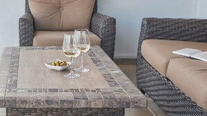 Un sofá de mimbre con un cojín color beige, una mesita con cubierta de mosaico, dos copas de vino y un bol de aceitunas.