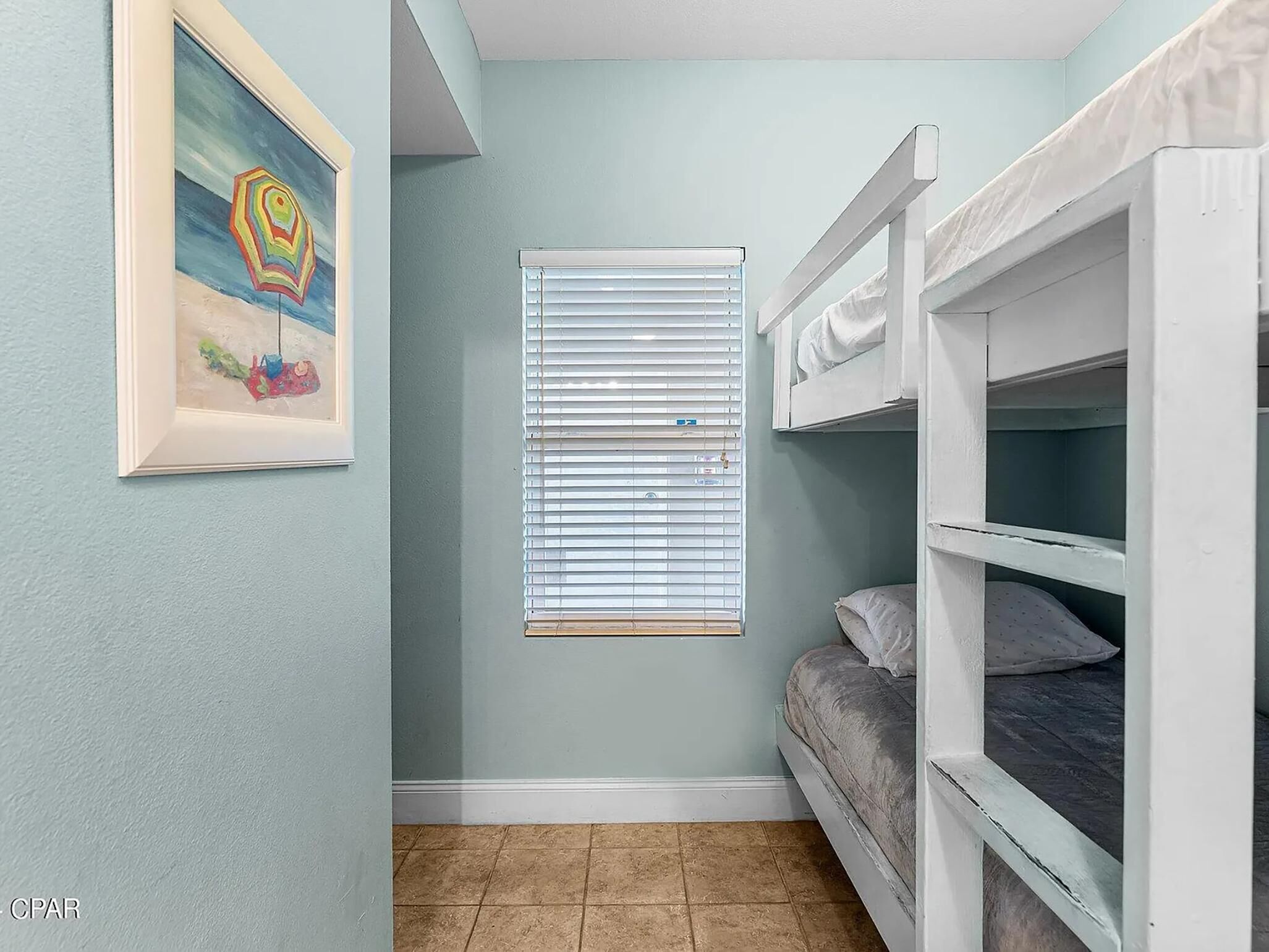 Une chambre d’enfant avec des lits superposés, une fenêtre avec des stores et un tableau encadré accroché au mur.