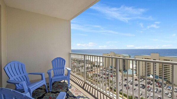 A balcony with two blue chairs, a patterned rug, and a view of a parking lot and ocean. - Laketown Wharf 1523 (Panama City Beach)