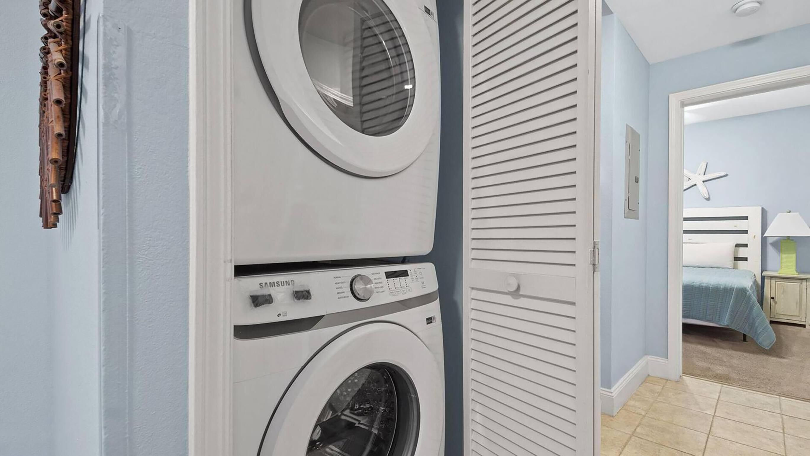 A laundry room with two stacked washing machines, a wooden door, and a view into a bedroom with a bed and a starfish decoration.