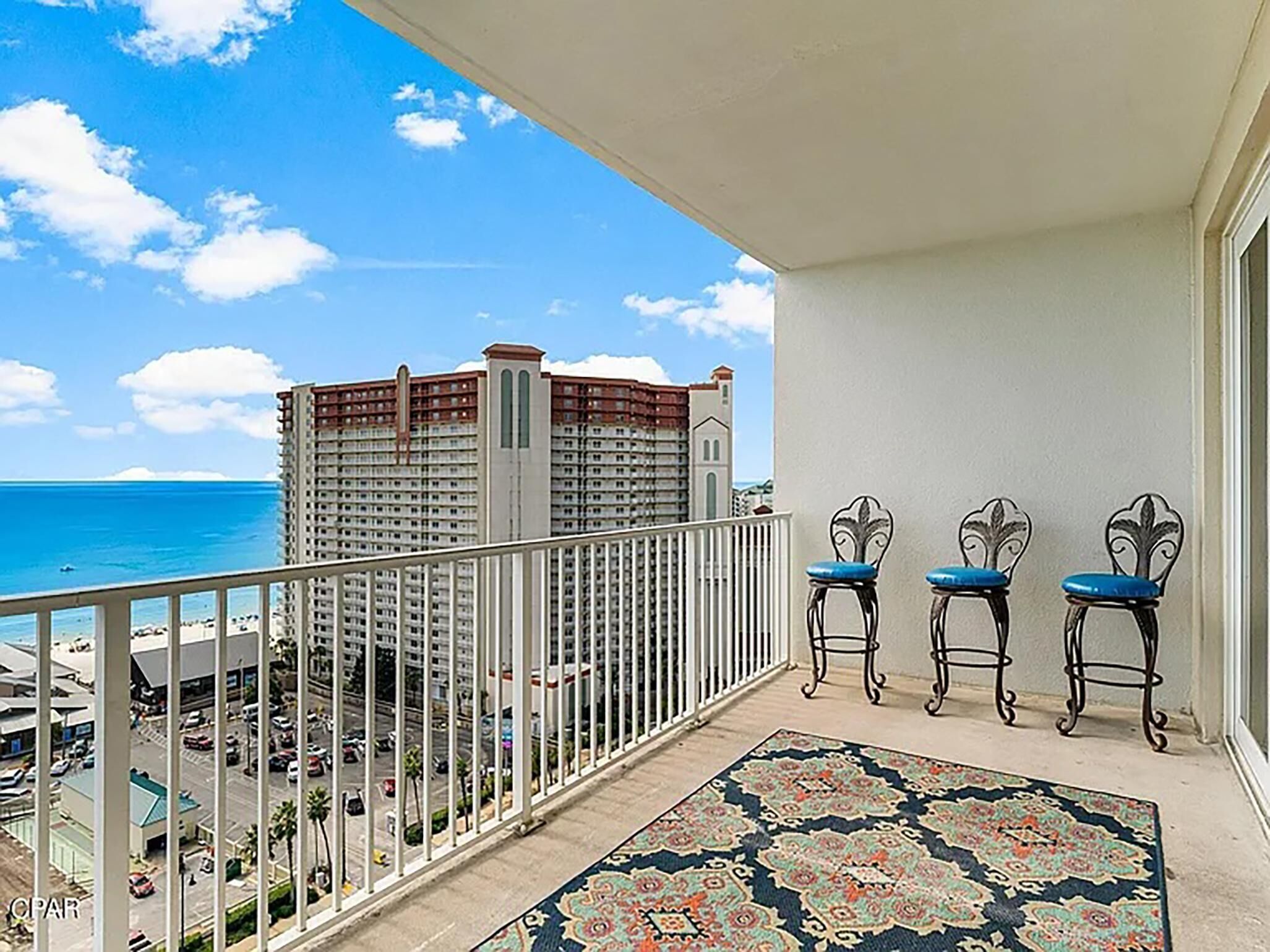 Un balcon avec vue sur l’océan et un immeuble de grande hauteur. On y trouve trois chaises avec des coussins bleus et un tapis à motifs sur le sol.