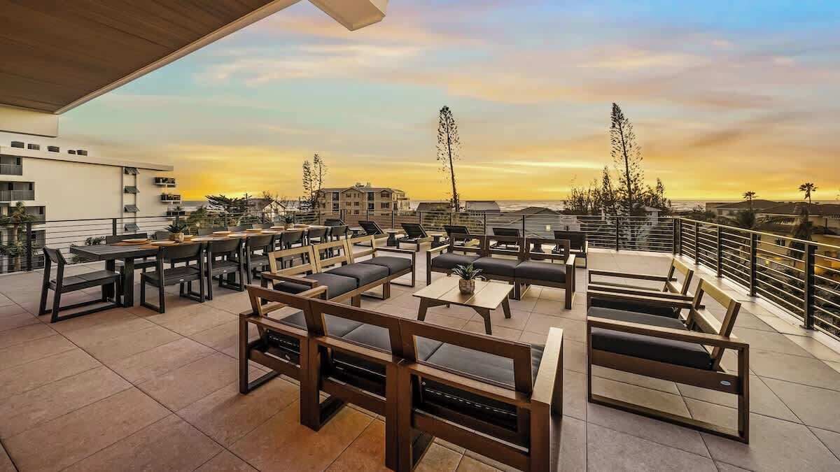 A rooftop terrace with outdoor seating, including tables and sofas, and a view of the sunset over the cityscape.