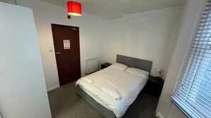 A bedroom with a bed, a nightstand, a red hanging light, a door, and a window with blinds. - Seaside Hotel  (Brighton)