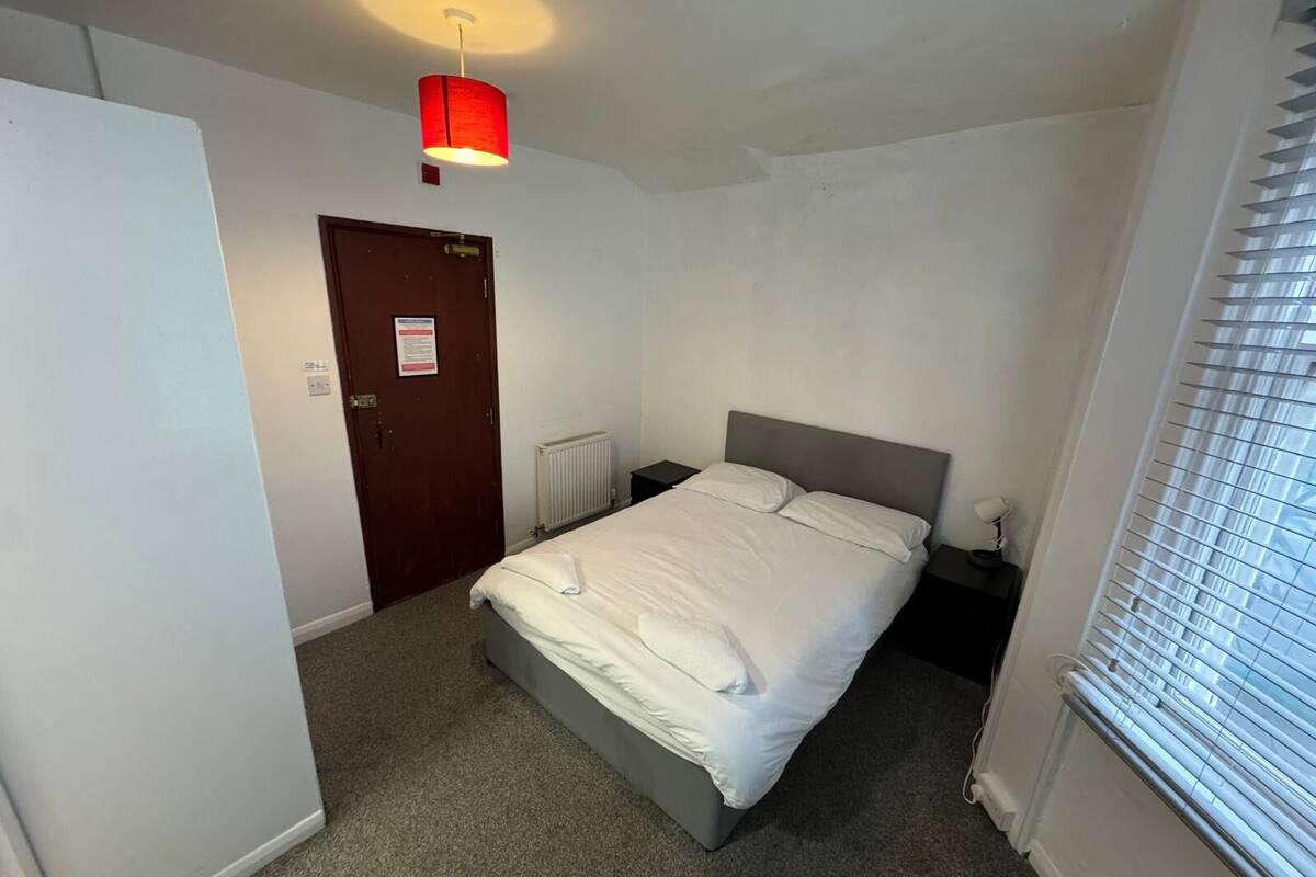 Un dormitorio con cama, mesita de noche, lámpara colgante roja, una puerta y una ventana con persianas.