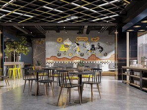 A modern restaurant interior with a high ceiling, geometric ceiling design, and a mural depicting cyclists. Tables and chairs are arranged for dining. - Yello Hotel Harbour Bay Batam (Batam)