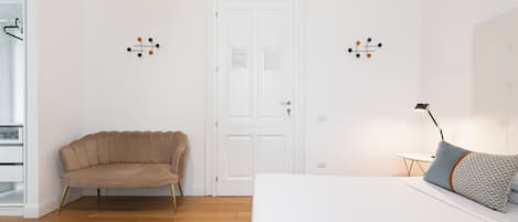 Une pièce avec un canapé beige, un lit blanc avec des coussins à motifs, une petite table avec une lampe et une porte blanche ornée d’un tableau d’affichage.