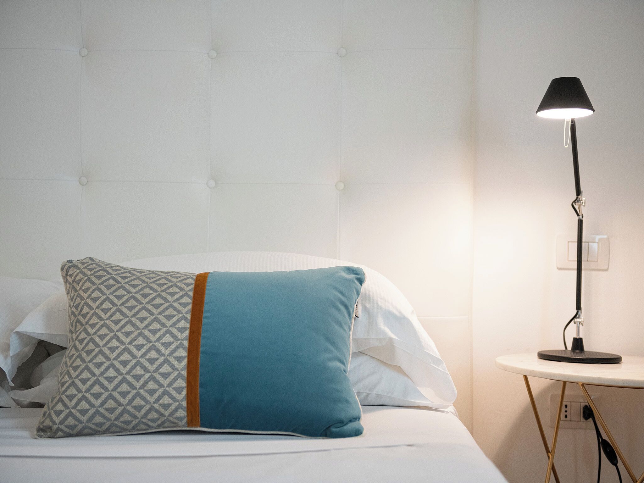 Un lit bien fait, agrémenté d’oreillers à motifs, une lampe de chevet et un mur blanc avec un interrupteur.