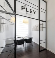 Un interno moderno con una parete divisoria in vetro con la scritta "PLEY HERITAGE", un tavolo da pranzo con sedie e una lampada a sospensione.