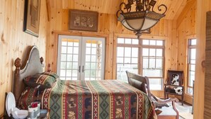 A wooden cabin bedroom with a bed, a chair, a table with a mirror, and a window with a view. - SKYLINE LODGE~ Spectacular Mountain Escape! (fletcher)