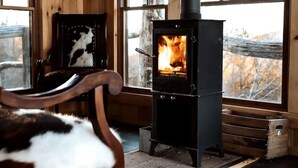 A cozy room with a wood-burning stove, a chair with a cow print cushion, and a window with a view of trees. - SKYLINE LODGE~ Spectacular Mountain Escape! (fletcher)