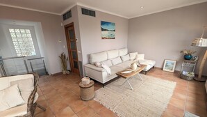 Een woonkamer met een bank, een salontafel en een rieten stoel. Er is een deur met een raam, een schilderij aan de muur en een mand op de vloer.