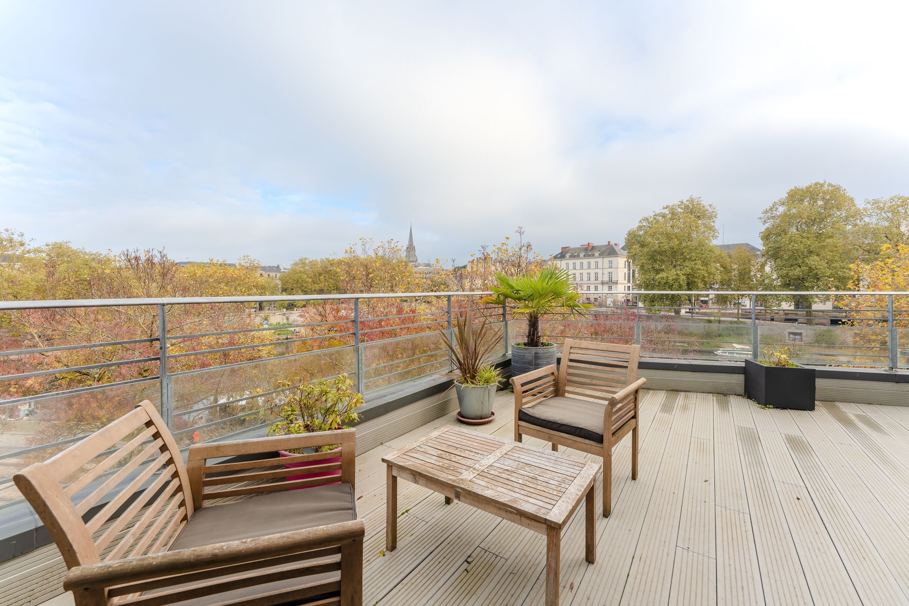 Une terrasse en bois avec une table et des chaises, des plantes en pot et une vue sur un paysage urbain, avec des arbres aux couleurs automnales et le clocher d’une église.