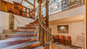 Un escalier doté d’une rampe en bois et d’une corde, menant à une pièce dont le sol est en pierre et le mobilier en bois.