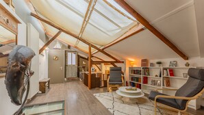 Un loft spacieux avec de hauts plafonds, des poutres apparentes, une lucarne, une grande bibliothèque, un fauteuil confortable et une table basse en verre.