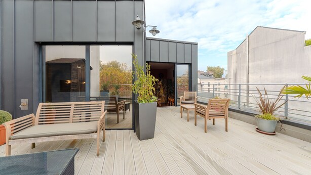 Une terrasse extérieure moderne, dotée de mobilier en bois, d’un grand bac à plantes et d’une porte vitrée donnant sur un espace intérieur.