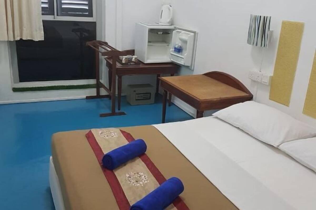 A hotel room with a bed, a desk, a chair, a small refrigerator, and a window with blinds.