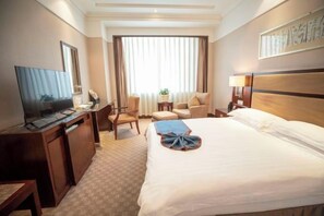 Habitación de hotel con cama, escritorio con televisor, silla, mesita de noche con lámpara y ventana con cortinas.