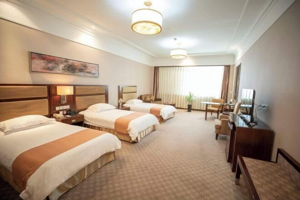 A hotel room with three beds, a desk, a television, and a large window with curtains.