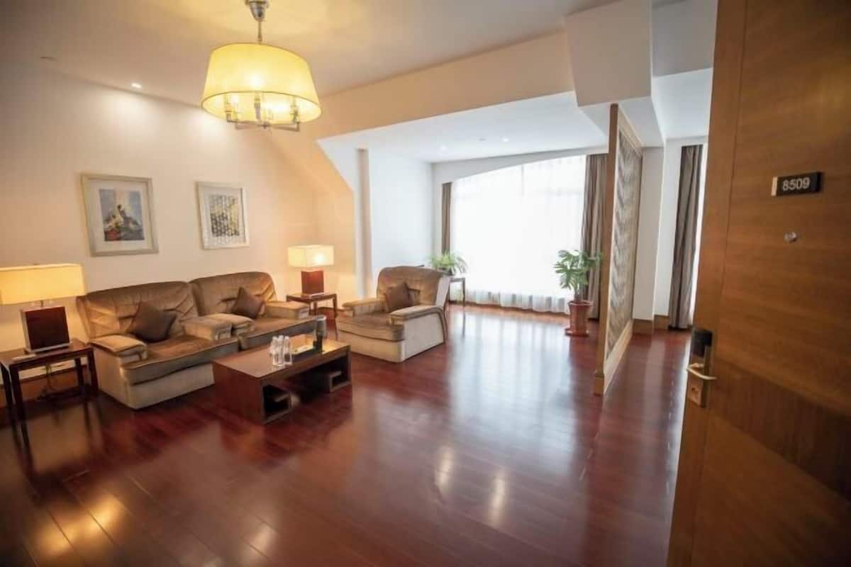 A spacious living room with a sofa set, a coffee table, and a lamp. There is a large window with curtains and a door with a number sign.