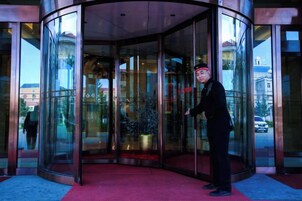 Una persona de pie frente a una puerta giratoria con paneles de vidrio, una alfombra roja y un edificio al fondo.