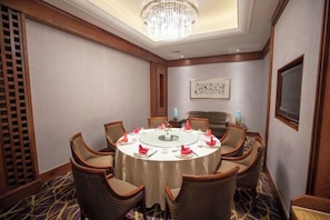 Une salle de conférence avec une table ronde, des chaises, un lustre et un téléviseur fixé au mur.