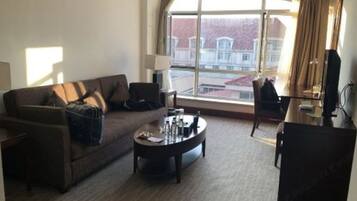 A living room with a sofa, coffee table, and a TV. There is a large window with a view of buildings outside.