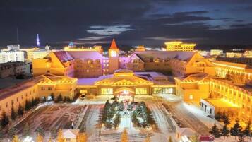 A large, illuminated building with a central tower and multiple wings, surrounded by snow-covered ground and trees.