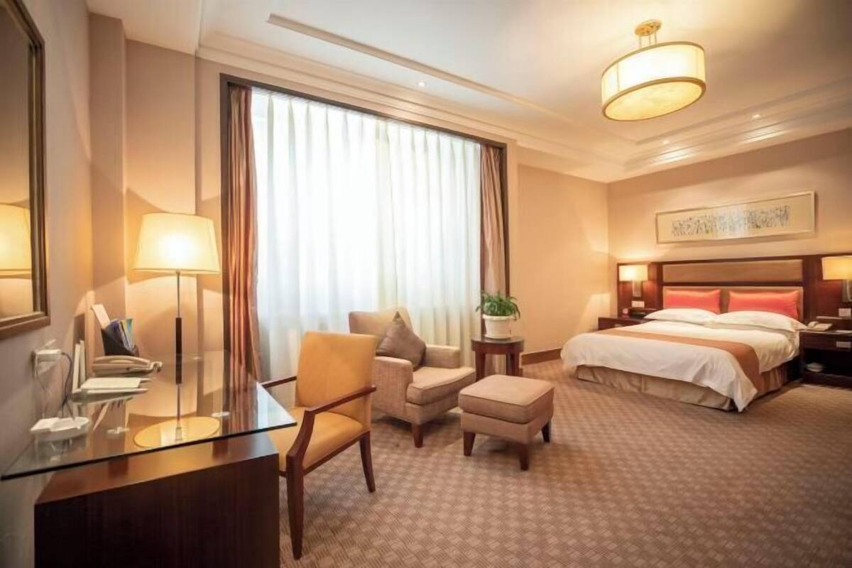 A hotel room with a bed, a desk, a chair, a small table with a plant, and a large window with curtains.