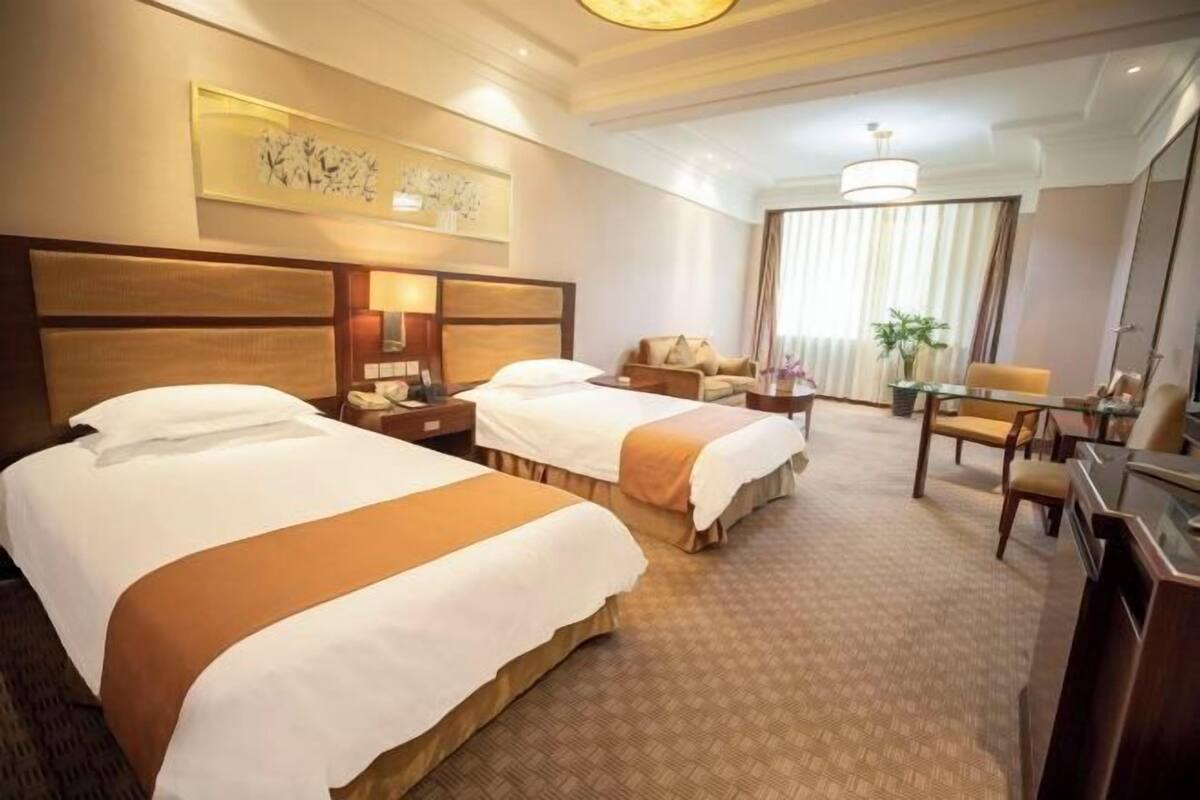 A hotel room with two beds, a desk, a chair, a sofa, a dining table, and a large window with curtains.