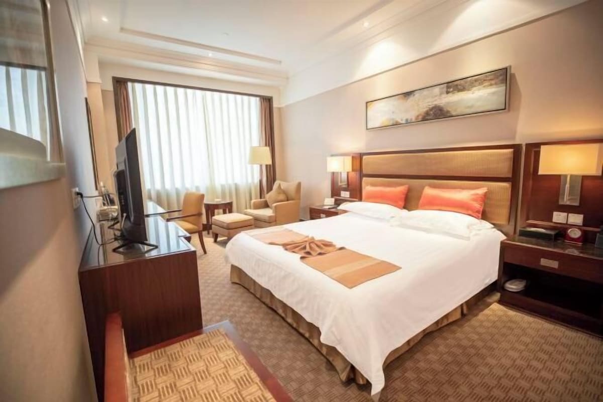 Habitación de hotel con una cama grande, un escritorio con televisor, una silla y una mesita de noche con lámparas.
