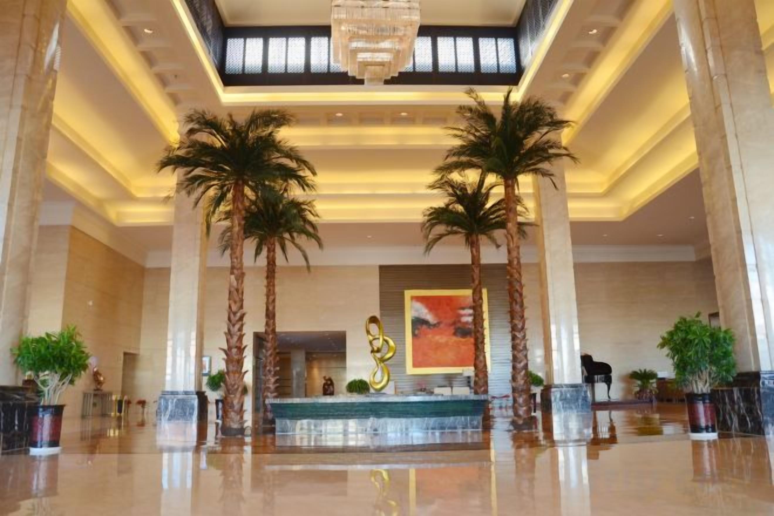 A spacious lobby with a reception desk, palm trees, and a modern chandelier.