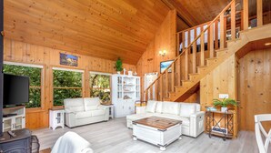 L’intérieur d’un chalet en bois comprend un salon, des canapés en cuir blanc, une table basse en bois, un téléviseur et un escalier.