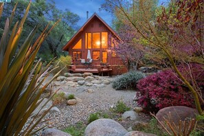 A wooden cabin with a deck, surrounded by rocks and plants.