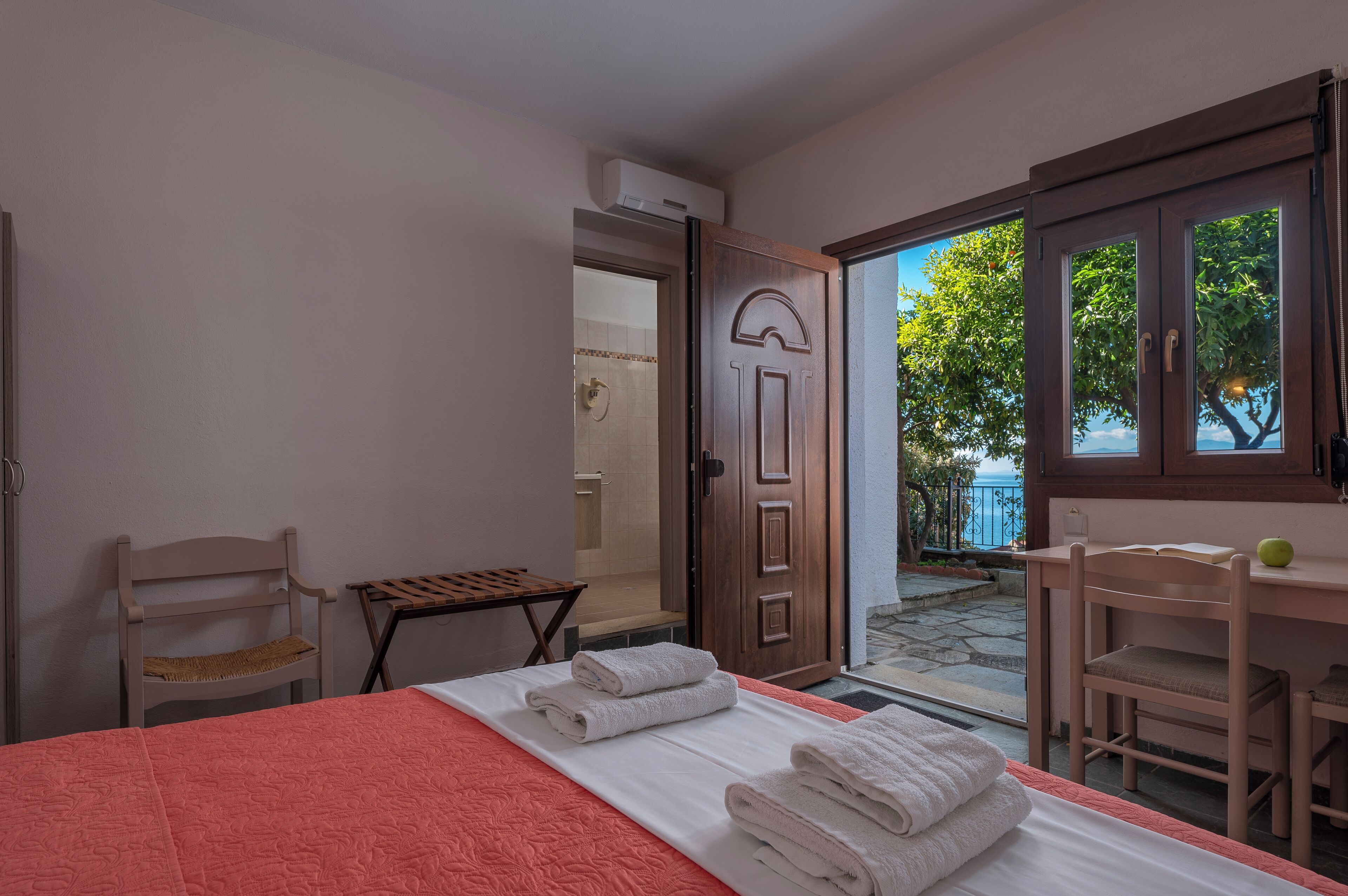 A hotel room with a bed, a wooden chair, a small table, and a door leading to an outdoor area with a view of trees and a railing.