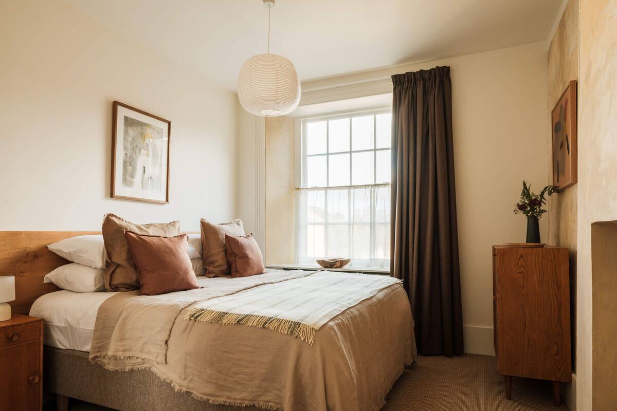 A neatly made bed with a wooden headboard, a bedside table, a hanging light fixture, a window with curtains, and a small wooden cabinet.