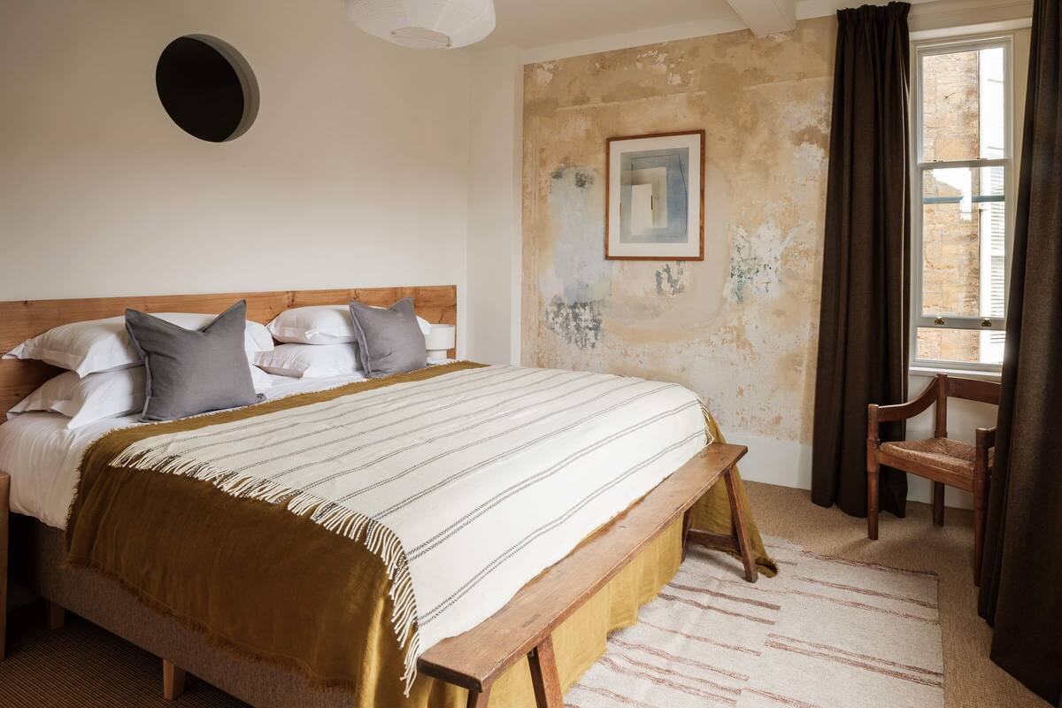 A bedroom with a large bed, a wooden headboard, a round wall light, a painting, and a window with curtains.