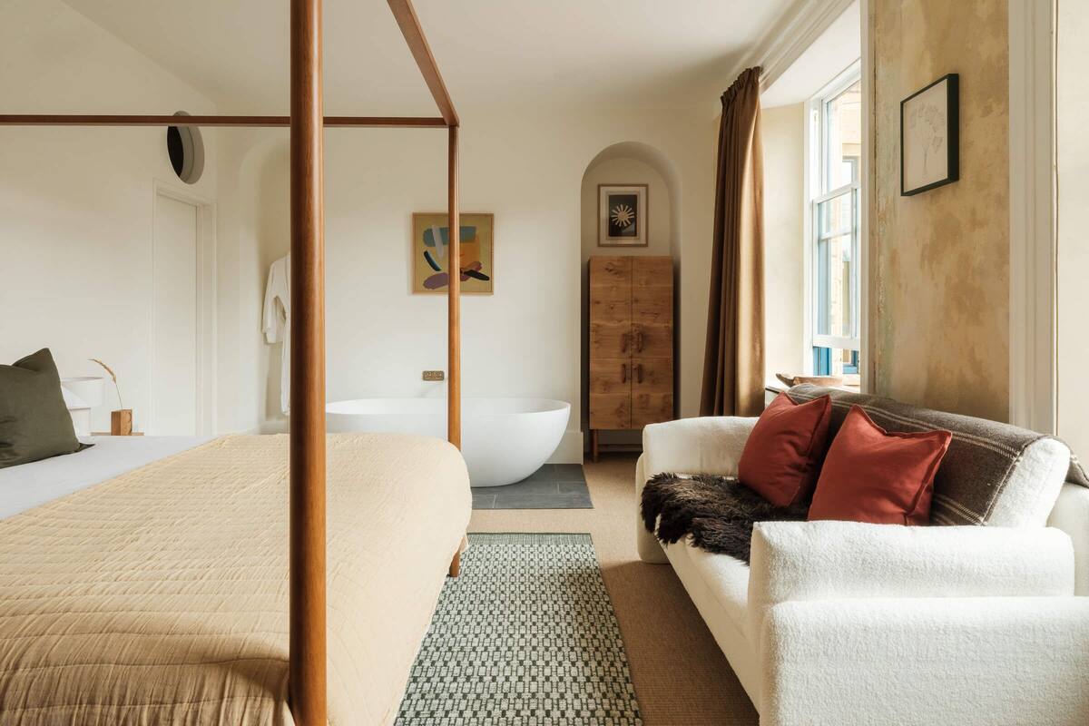 A bedroom with a four-poster bed, a white sofa, a wooden cabinet, and a large window.