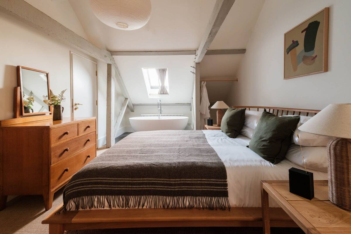 A bedroom with a large bed, a wooden dresser, a bedside table, a hanging light fixture, a window, and a painting on the wall.
