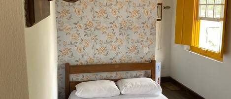 A bedroom with a wooden bed, floral wallpaper, a bedside lamp, and a window with yellow shutters.