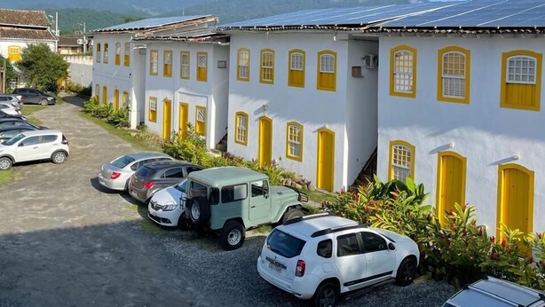 A row of white buildings with yellow doors and windows, parked cars, and mountains in the background. - Pousada Fortaleza (Paraty)