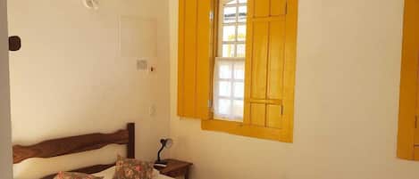 Une chambre à coucher comprenant un lit, un banc, une fenêtre avec des volets jaunes, un climatiseur et un ventilateur de plafond.