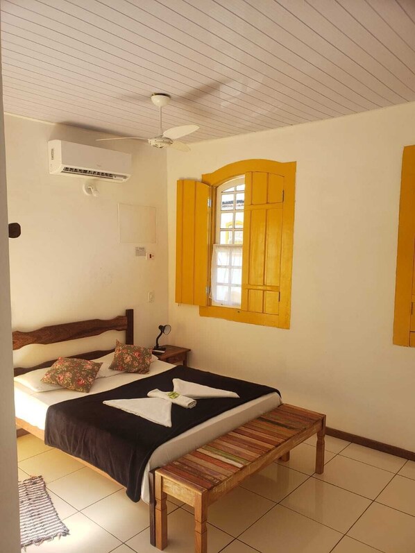 Un dormitorio con una cama, un banco, una ventana con contraventanas amarillas, un equipo de aire acondicionado y un ventilador de techo.